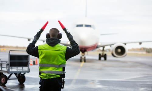 A runway marshall waving light wands to guide an airplane in front of them.