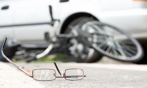 A pair of cracked eyeglasses lying on the road with a collision between a bicycle and a sedan in the background.