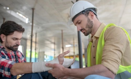 A hardhat employee assisting an injured coworker by wrapping gauze around his hand.