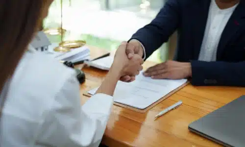 A personal injury lawyer seated behind a desk and shaking hands with her client as they are about to discuss a case.